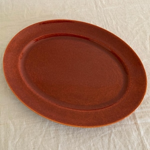 May include: An oval-shaped, reddish-brown ceramic platter. The serving dish has a slightly textured surface and a raised rim. The plate is empty and sits on a light-colored, textured surface.