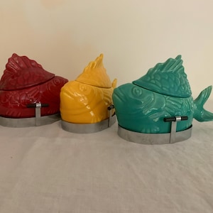 May include: Three ceramic fish-shaped lidded containers in red, yellow, and teal. Each container features a textured scale pattern and is held by a silver-toned metal stand with a small black handle detail on the front of the base.