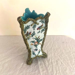 May include: A decorative vase with a light blue interior and a textured exterior. The vase features a detailed design of birds, flowers, and foliage on a white background, framed by a bronze-colored trim. The vase has a unique, irregular shape.