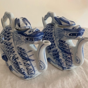 May include: Two matching white and blue ceramic teapots. Each teapot is shaped like a dragon with intricate blue detailing. The teapots have a handle, a spout, and a lid. The design is reminiscent of traditional Asian pottery.