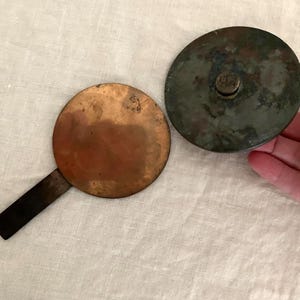 May include: A circular copper-colored metal disc with a rectangular handle attached to one side, positioned next to a circular, dark, weathered metal lid featuring a small central knob. Both items exhibit signs of age, oxidation, and patina.