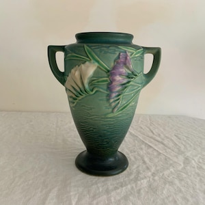 May include: A green ceramic vase with two handles and a wide base. The vase features embossed floral designs in white and purple. The vase has a textured surface and a dark green rim and base. The vase is approximately 10 inches tall.