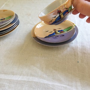 May include: A set of miniature teacups and saucers with a lighthouse and seascape design. The cups and saucers are painted in shades of yellow, blue, and green.