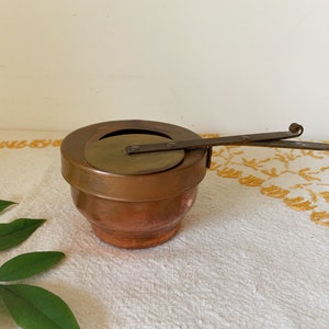 May include: A copper and brass warming pot with a hinged lid and a metal handle. The pot is sitting on a white tablecloth with a floral pattern.