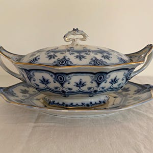 May include: A white porcelain serving dish with a blue and gold floral pattern. The dish has a lid and a matching base.