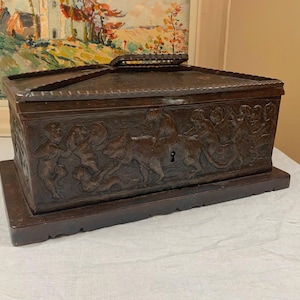 May include: A dark brown, rectangular wooden box with intricate carvings depicting figures and animals. The box has a hinged lid with a handle and sits on a matching base. A keyhole is visible on the front.