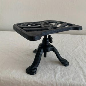 May include: A black cast iron trivet with a decorative floral design on the square top. The trivet has three curved legs and an adjustable height mechanism. This kitchenware item is designed to protect surfaces from hot cookware.