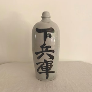 May include: A tall, light gray ceramic bottle with a narrow neck and rounded body. The bottle features black Japanese characters running vertically down the front. The bottle is sitting on a white surface.