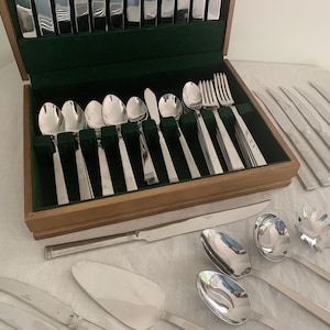 May include: A wooden cutlery box with a green velvet interior holds various stainless steel spoons, forks, and knives. Additional silverware, including serving spoons and a cake server, is arranged around the box on a white surface.