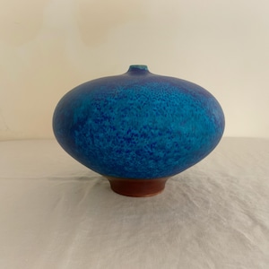 May include: A rounded ceramic vase with a wide, bulbous body featuring a mottled blue glaze that transitions into darker tones. The vase has a small, narrow opening at the top and sits on a solid, unglazed brown circular base.