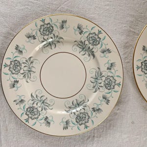 May include: Two off-white plates with a floral design in teal and gray, accented with gold rims. The plates feature a central circle and a repeating pattern of stylized flowers and leaves around the edges. A classic dinnerware set.