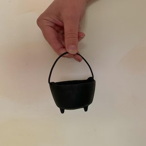 May include: A small, black cast iron cauldron with a curved handle. The cauldron has three small feet and a rounded body. The handle is made of a thin, black material. The cauldron is held by a hand.