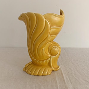 May include: A yellow ceramic vase with a seashell design. The vase has a curved, open top and a detailed, textured surface. The base is a rounded shape with a scalloped edge. The vase is approximately 10 inches tall.