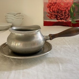 May include: A small, antique pewter saucepan with a dark wooden handle, resting on a matching saucer. The pot has a rounded body and a pouring spout. The handle is attached with metal rivets. The set is displayed on a white tablecloth.