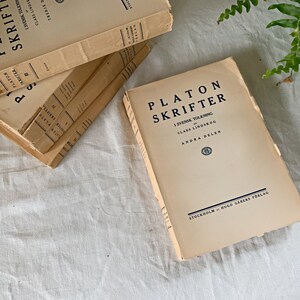 May include: A stack of beige books with the title "Platon Skrifter" in black text. The top book has the text "I Svensk Tolkning" and "Clas Lindskog" in smaller black text. The bottom book has the text "Andra Delen" and "Stockholm Hugo Gebers Forlag" in smaller black text.