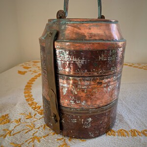 May include: A three-tiered copper lunch box with a brass handle and clasps. The box has a worn, aged patina and is sitting on a white tablecloth with a floral pattern.
