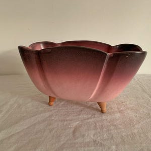 May include: A vintage, petal-shaped planter with a gradient color scheme, transitioning from a deep burgundy at the rim to a soft pink at the base. The ceramic planter rests on three small, wooden legs, adding a touch of mid-century charm.
