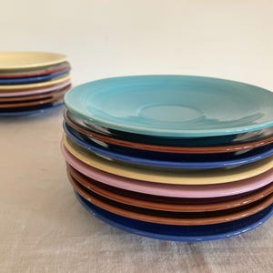 May include: Stacks of colorful ceramic plates in various shades, including blue, brown, pink, and yellow. The plates are arranged in two separate stacks, showcasing their vibrant colors and glossy finish. A simple, elegant design.