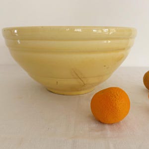 May include: A large, pale yellow ceramic mixing bowl with horizontal ridges. Three oranges are scattered in front of the bowl on a white surface. The bowl has a wide opening and a rounded base.