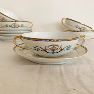 May include: A set of white porcelain soup bowls and saucers with gold trim and handles. The bowls and saucers feature a decorative floral and scroll design in pink, blue, and gold. Several sets are stacked.