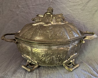 May include: An antique silver serving dish with a lid. The dish is decorated with intricate floral and scrollwork designs. The lid features a sculpted figure of a man holding a staff.