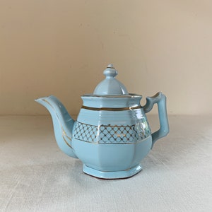 May include: A light blue teapot with a gold trim and a geometric pattern. The teapot has a lid and a handle.