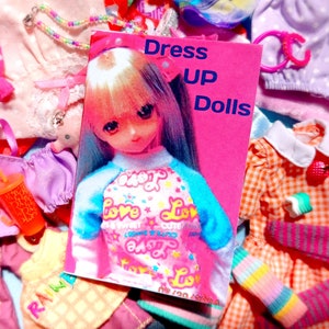 Op de afbeelding: Een roze boek met de titel "Dress Up Dolls" met een pop die een blauw en roze shirt draagt. De afbeelding is omgeven door poppenkleding en accessoires in verschillende kleuren, waaronder roze, oranje en paars. Een speelgoedbeker is ook zichtbaar.