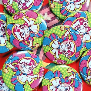 May include: A collection of round, colorful buttons featuring a cartoon jester character. The jester has a pink and blue harlequin hat and outfit, with a green polka dot background. The buttons are arranged on a red surface.