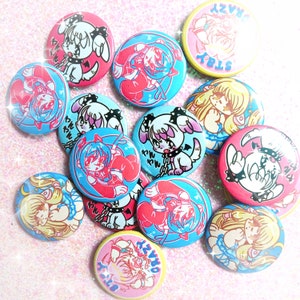 May include: A collection of 12 colorful round pins featuring cartoon characters and Japanese text. The pins are decorated with various designs, including animals, people, and hearts. The pins are all different colors, including pink, blue, yellow, and red.