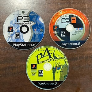 May include: Three PlayStation 2 video game discs. The discs feature the titles "Persona 3" and "Persona 4" with character art and logos. The discs are on a wooden surface.