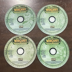 May include: Four World of Warcraft: Burning Crusade game discs. Each disc is green with the game title and disc number. The discs are arranged on a brown surface.