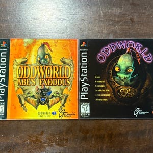 May include: Two PlayStation game cases, "Oddworld: Abe's Exoddus" and "Oddworld: Abe's Oddysee." The "Abe's Exoddus" case has an orange background with a green alien. The "Oddysee" case is black with a green alien.
