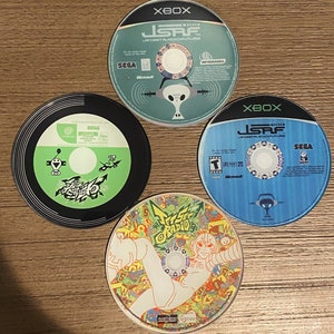 May include: Four video game discs are arranged on a wooden surface. Two discs are labeled "XBOX" and "JSRF JET SET RADIO FUTURE". One disc is black and green with "JET SET RADIO" text. The fourth disc has a colorful graphic.