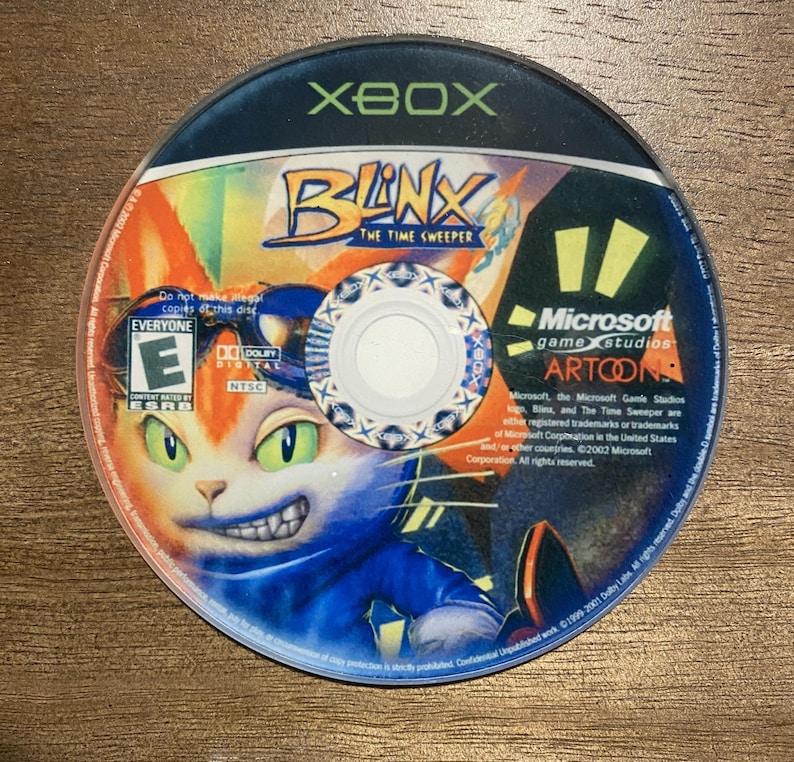 May include: A circular Xbox game disc for "Blink: The Time Sweeper." The disc features a colourful illustration of a cat-like character with green eyes and a blue outfit. The disc also displays the Xbox logo, ESRB rating, and Microsoft Game Studios branding.