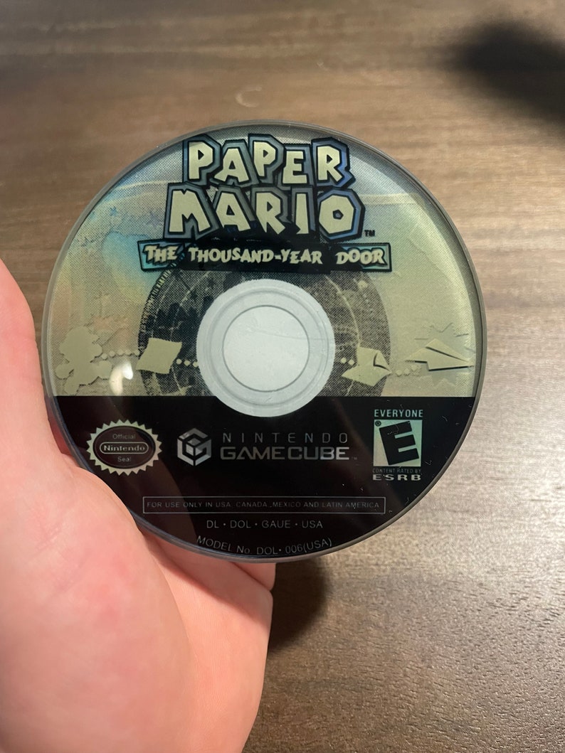 May include: A GameCube disc for Paper Mario: The Thousand-Year Door. The disc has the game's title in blue and white. The Nintendo GameCube logo and the ESRB rating are also visible on the disc.