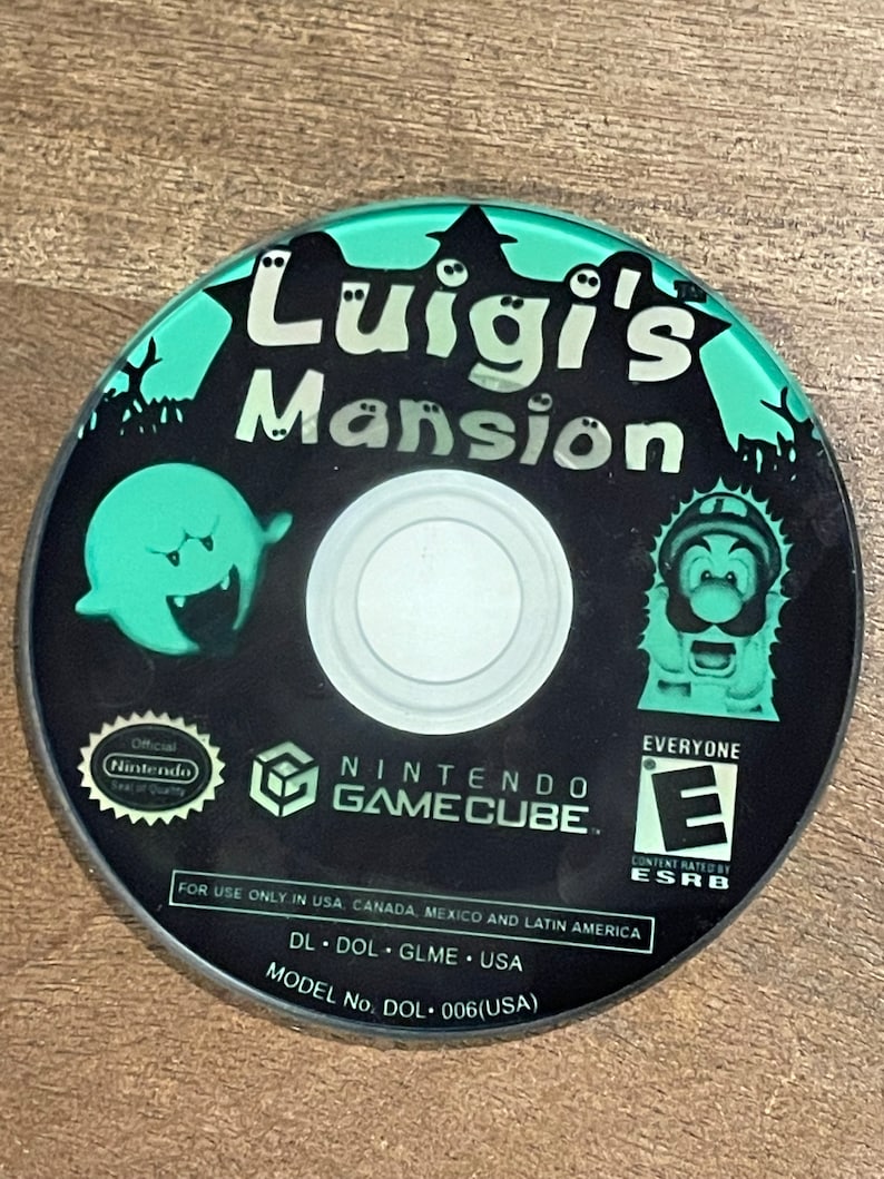 May include: A black Nintendo GameCube disc for "Luigi's Mansion" with a teal and white design. The disc features a ghost, Luigi, and the game's title. The ESRB rating is "E" for Everyone.