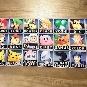 May include: A collection of rectangular stickers featuring characters from the Super Smash Bros. video game series. Each sticker displays a character's portrait and name, including Mario, Luigi, and Pikachu. The stickers are arranged on a wooden surface.