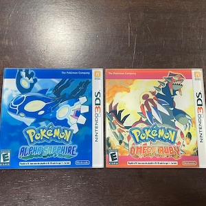 May include: Two Nintendo 3DS game cases: Pokémon Alpha Sapphire and Omega Ruby. The Alpha Sapphire case is blue with a white and blue creature graphic. The Omega Ruby case is red with a white and red creature graphic. Both cases have the Nintendo 3DS logo.