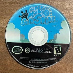 May include: A Super Mario Sunshine Nintendo GameCube disc. The disc features a blue sky background with the game title in white and blue. The disc also includes the Nintendo GameCube logo and ESRB rating.