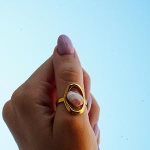 Puede incluir: Un anillo de tono dorado con un marco ovalado que rodea una pequeña concha marina lisa con rayas blancas y marrones. La banda del anillo es de metal sólido de tono dorado y el engaste central mantiene la concha natural firmemente en su lugar.