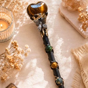 K&ouml;nnte beinhalten: Ein schwarzer Zauberstab mit einem herzf&ouml;rmigen braunen Edelstein an der Spitze, akzentuiert durch ein silberfarbenes Schmetterlingsornament. Der Stab ist mit Metalldraht umwickelt und mit drei kugelf&ouml;rmigen Edelsteinen in Gr&uuml;n, Braun und Blaugrau verziert.