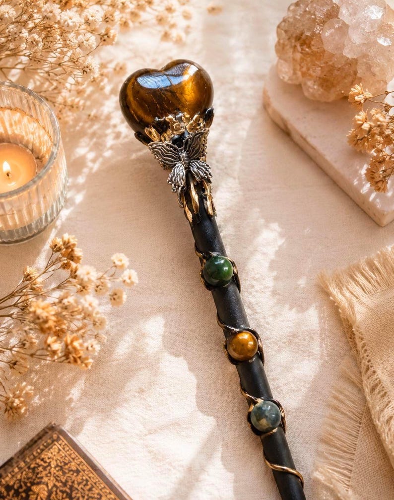 K&ouml;nnte beinhalten: Ein schwarzer Zauberstab mit einem herzf&ouml;rmigen braunen Edelstein an der Spitze, akzentuiert durch ein silberfarbenes Schmetterlingsornament. Der Stab ist mit Metalldraht umwickelt und mit drei kugelf&ouml;rmigen Edelsteinen in Gr&uuml;n, Braun und Blaugrau verziert.