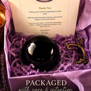 May include: A black polished sphere sits in a purple paper-filled box alongside a small ornate gold-toned metal stand. A thank you note from Dream Shop Crafting Co. is included, with text stating: PACKAGED with care & intention. EACH ORDER IS PREPARED.