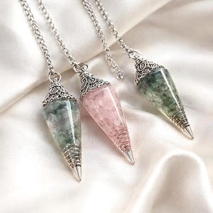 Op de afbeelding: Drie kristallen pendels met zilveren kettingen en sierlijke doppen. Eén is roze, één is groen en één is een mix van groen en wit. De pendels zijn kegelvormig met een zilveren punt, tentoongesteld op een witte, zijdeachtige ondergrond.