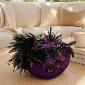 Può includere: Un oggetto decorativo arrotondato in velluto viola ornato da un denso grappolo di piume nere e piume più piccole con punte viola. La consistenza è morbida e soffice, caratterizzata da una tonalità viola intenso contrastata dal piumaggio scuro.
