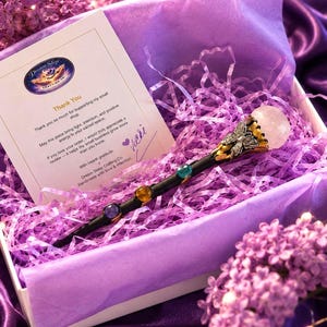 K&ouml;nnte beinhalten: Ein schwarzer Zauberstab mit silbernem Schmetterlingsakzent und einer runden, klaren Kristallspitze, besetzt mit drei bunten Steinen. Er liegt in einer wei&szlig;en Schachtel mit violettem F&uuml;llpapier neben einer Dankeskarte von Dream Sleep Crafting Co.