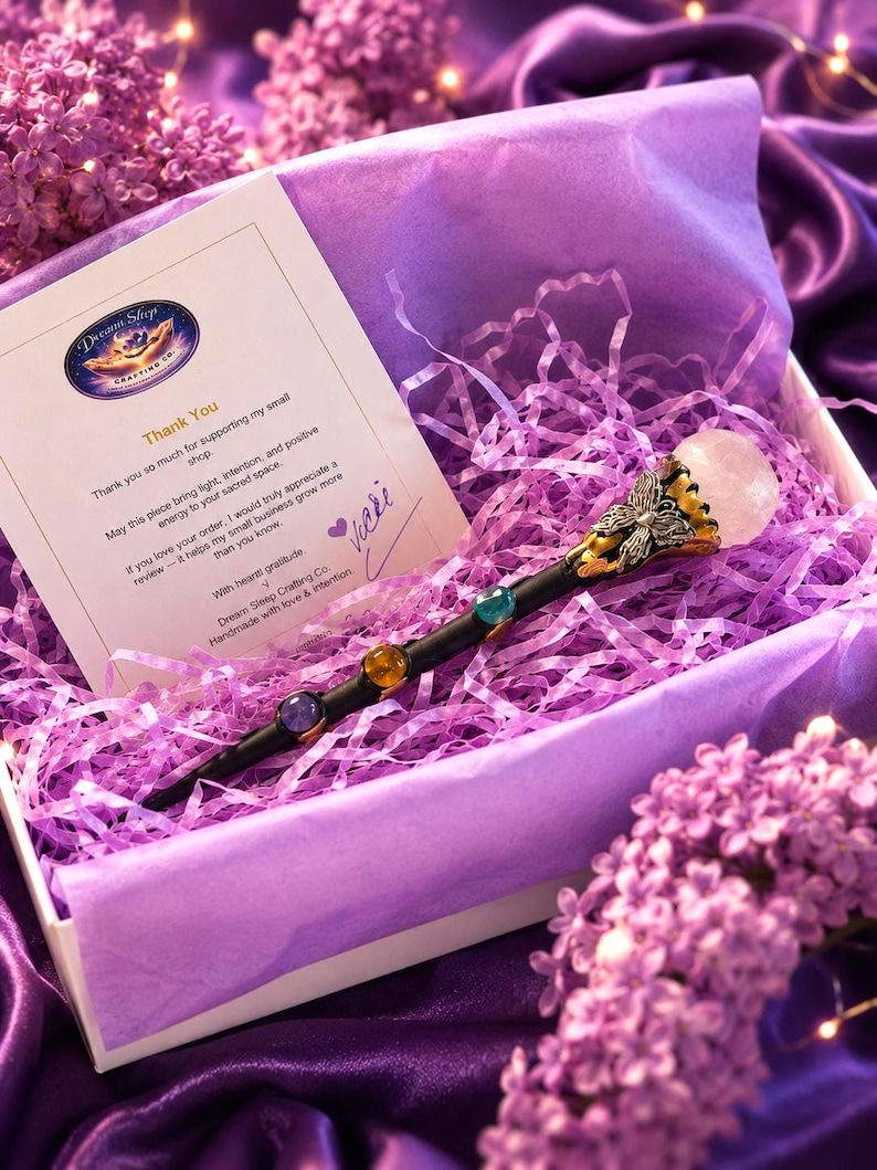 K&ouml;nnte beinhalten: Ein schwarzer Zauberstab mit silbernem Schmetterlingsakzent und einer runden, klaren Kristallspitze, besetzt mit drei bunten Steinen. Er liegt in einer wei&szlig;en Schachtel mit violettem F&uuml;llpapier neben einer Dankeskarte von Dream Sleep Crafting Co.