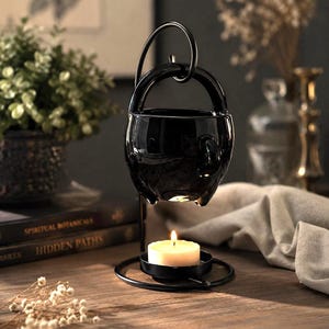May include: A black ceramic cauldron-shaped oil warmer with a handle, suspended above a black metal stand holding a lit candle. The candle's warm glow illuminates the space, creating a cozy ambiance. The background includes books and decorative elements.