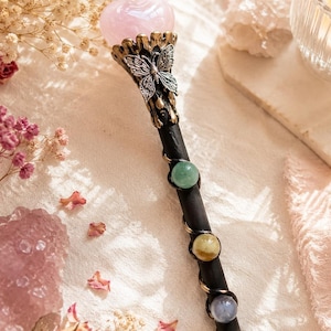 K&ouml;nnte beinhalten: Ein schwarzer Zauberstab mit einem herzf&ouml;rmigen, hellrosa Stein an der Spitze, der von einer silberfarbenen Schmetterlingsfassung gehalten wird. Der Stab ist mit einem d&uuml;nnen Metalldraht umwickelt, der drei kleine, runde, bunte Steine h&auml;lt.