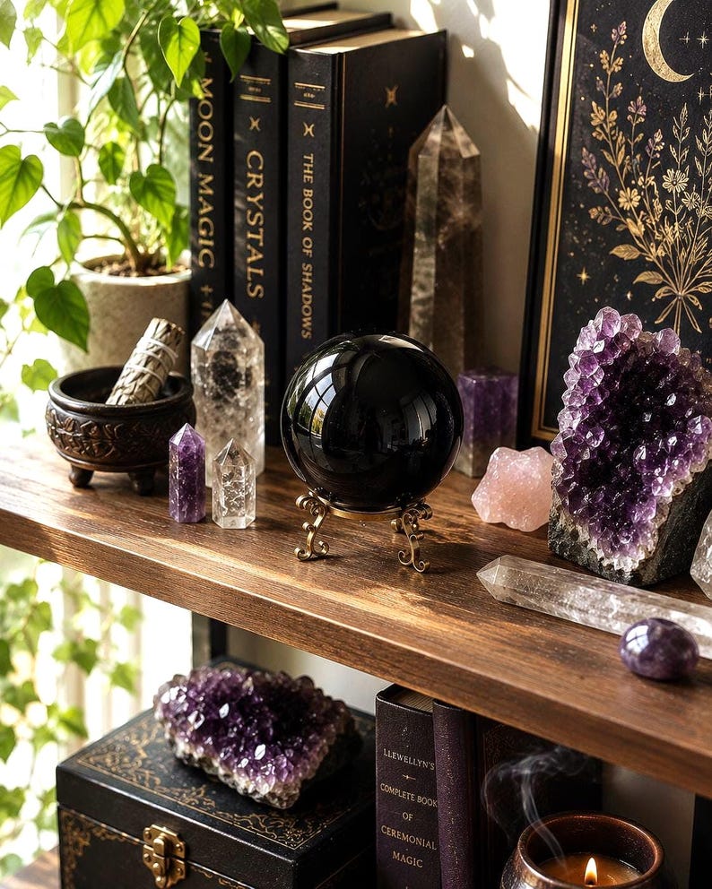 May include: A polished black obsidian sphere resting on a decorative gold-toned metal stand, surrounded by various crystals including amethyst clusters, clear quartz points, and a rose quartz piece, alongside black books and a small incense bowl.